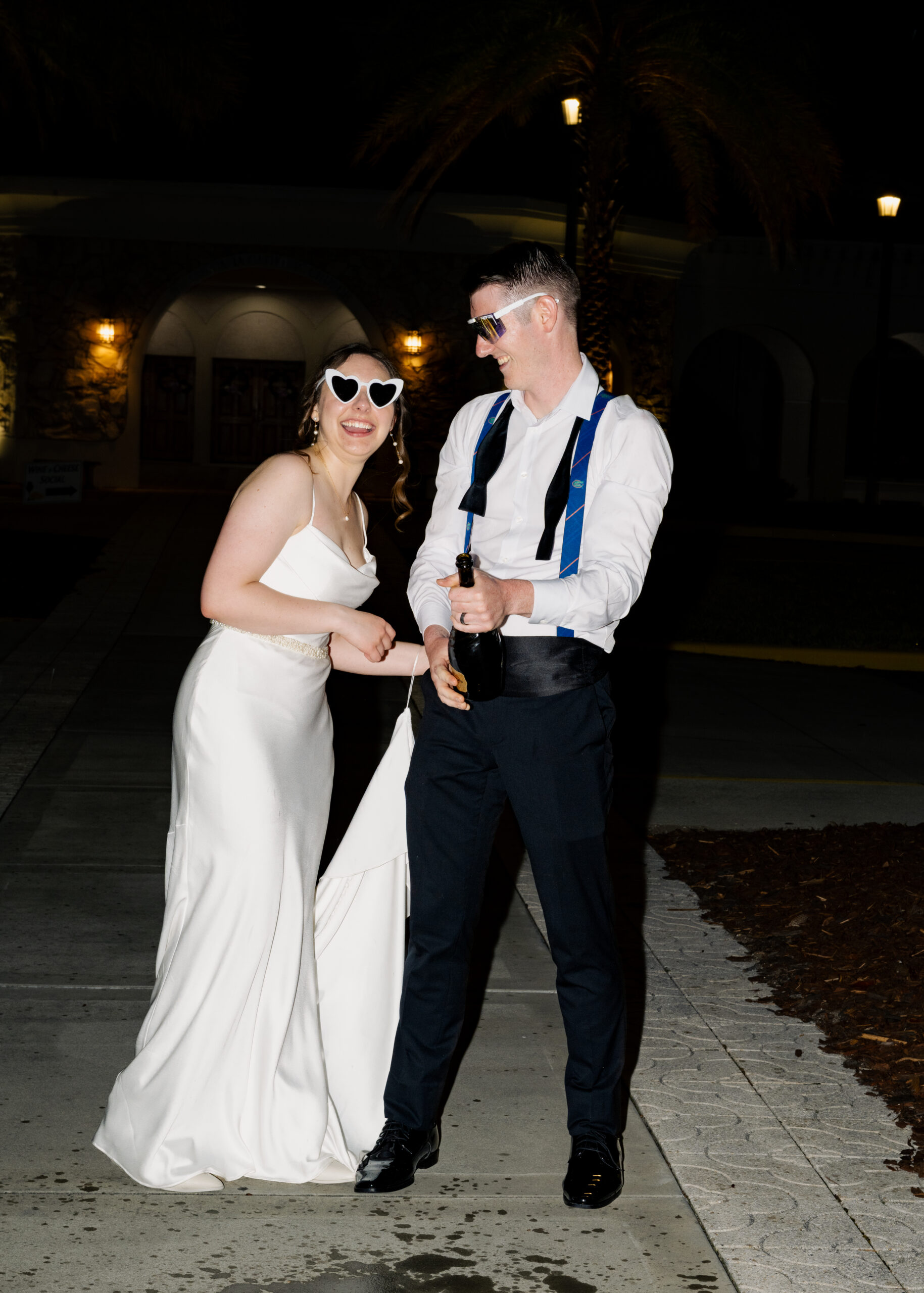Laughing bride and groom celebrate with a champagne bottle during their nighttime wedding reception portraits at St. Anastasia Catholic Church in St. Augustine, FL.
