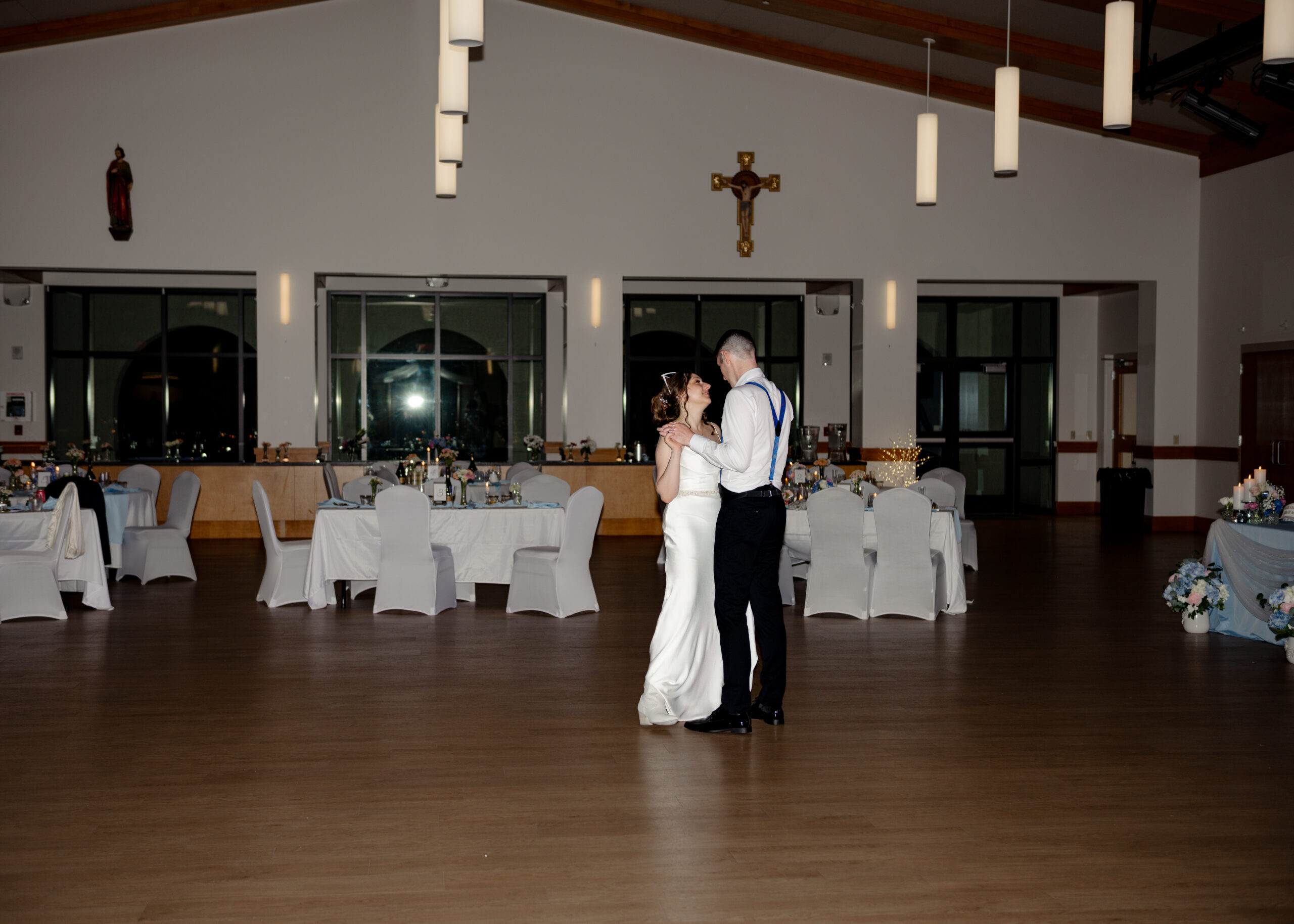 Bride and groom share a romantic last dance in the center of a spacious Catholic parish hallat st. anastasia catholic church in st augustine, florida, with elegant white-covered tables and soft overhead lighting in the background.