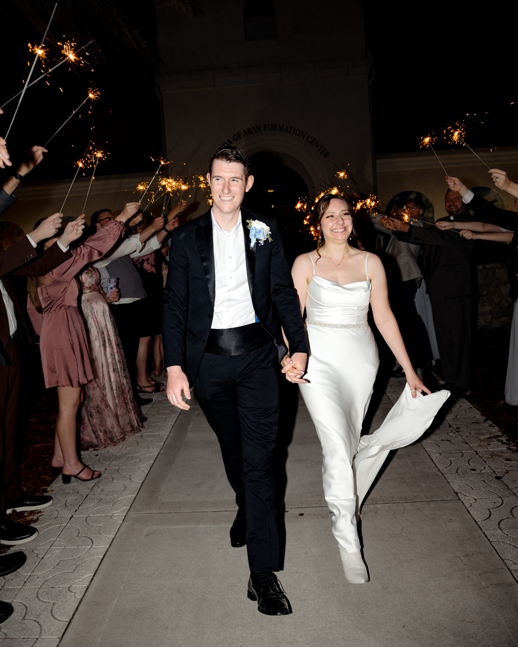 The bride and groom walk through a festive sparkler tunnel held by cheering guests, exiting their Catholic wedding reception at st. anastasia catholic church in st. augustine, florida with joyful expressions and elegance.