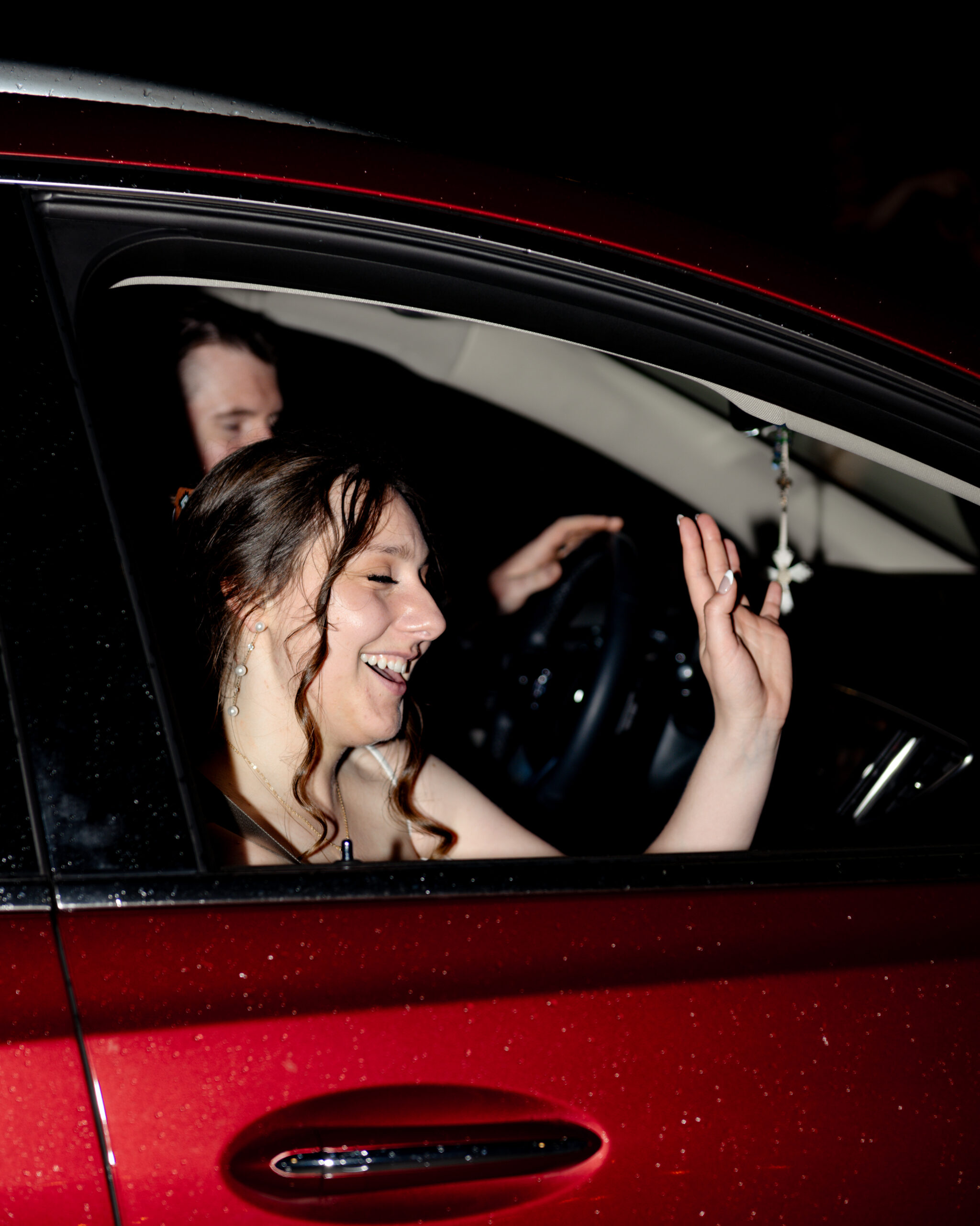 The bride waves joyfully from the passenger seat of a red car during the couple's grand departure after their Catholic wedding celebration.