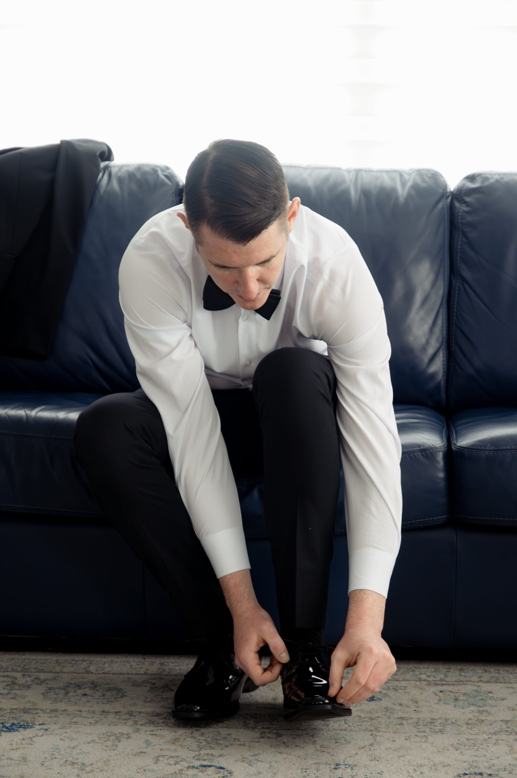 The groom tying his shoes while getting ready for his Catholic wedding in St. Augustine, Florida. Dressed in a classic white dress shirt and black bow tie, he prepares for the big day, showcasing the groom's attention to detail in his attire.