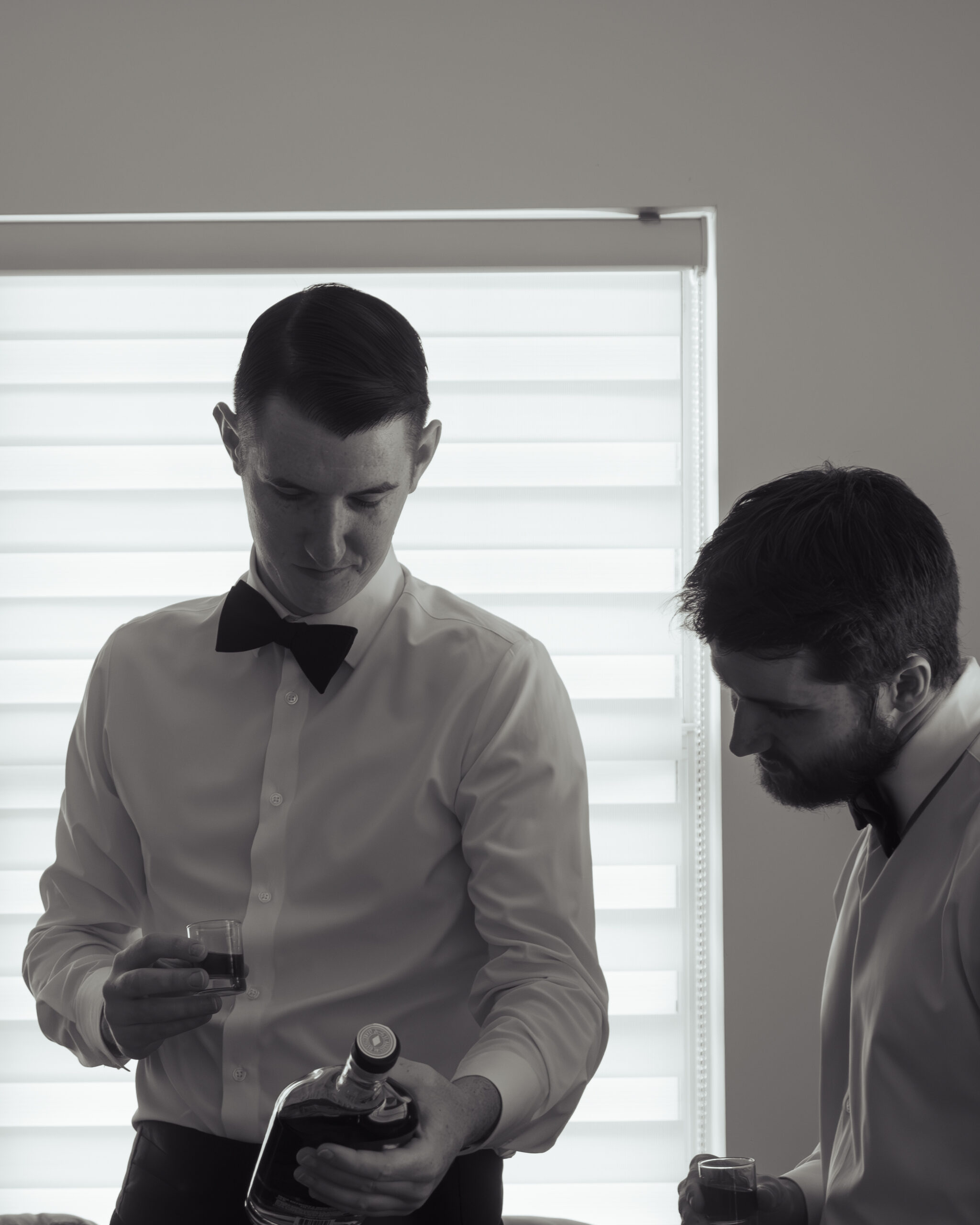 A black-and-white photo of the groom and a friend enjoying a moment while getting ready for his Catholic wedding in St. Augustine, Florida. The groom is holding a glass of whiskey, adding a personal touch to the pre-wedding preparations.
