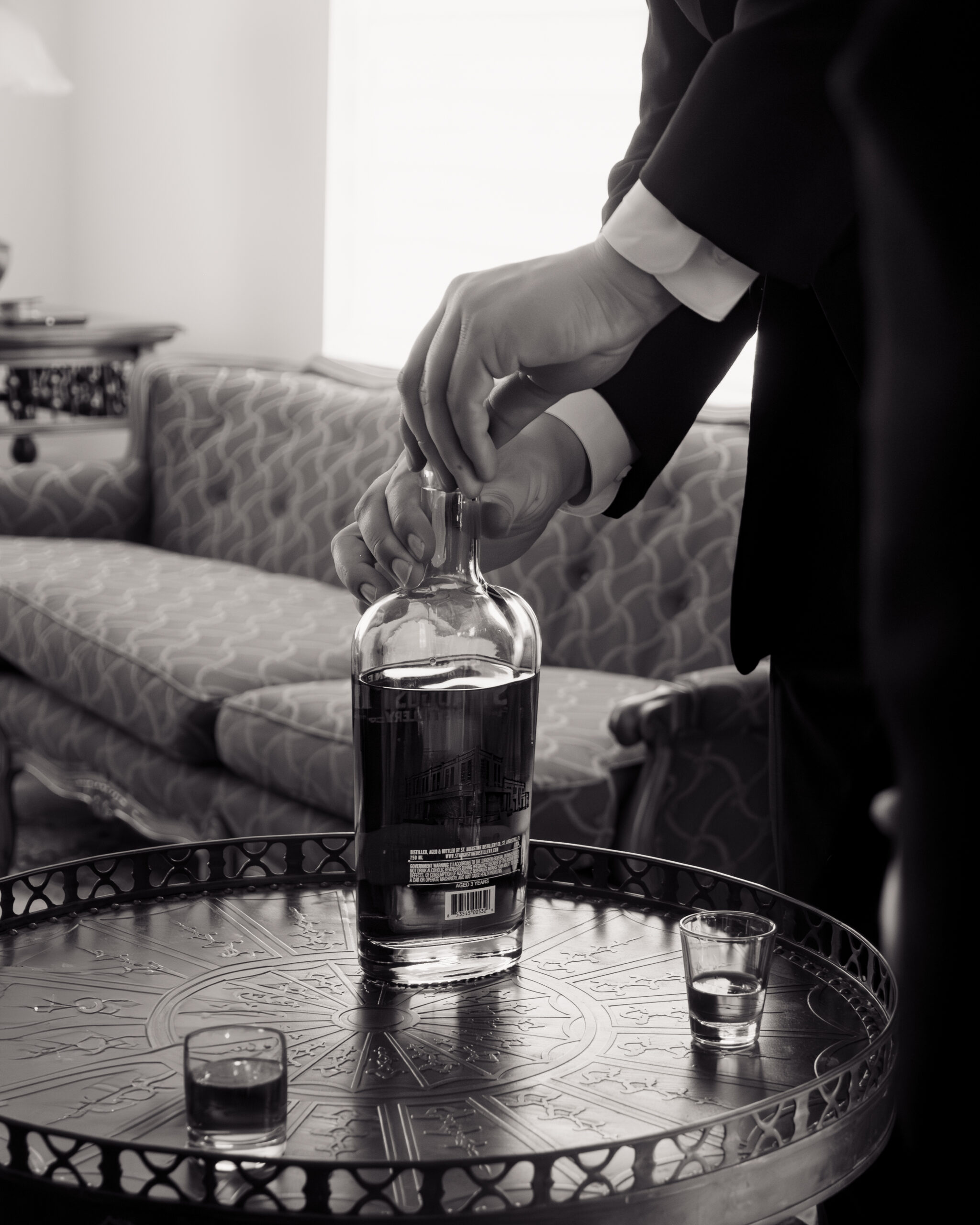 A close-up black-and-white shot of the groom pouring a drink from a bottle, preparing for his Catholic wedding in St. Augustine, Florida. The details of the bottle and glasses add a personal, intimate element to the groom's pre-wedding ritual.