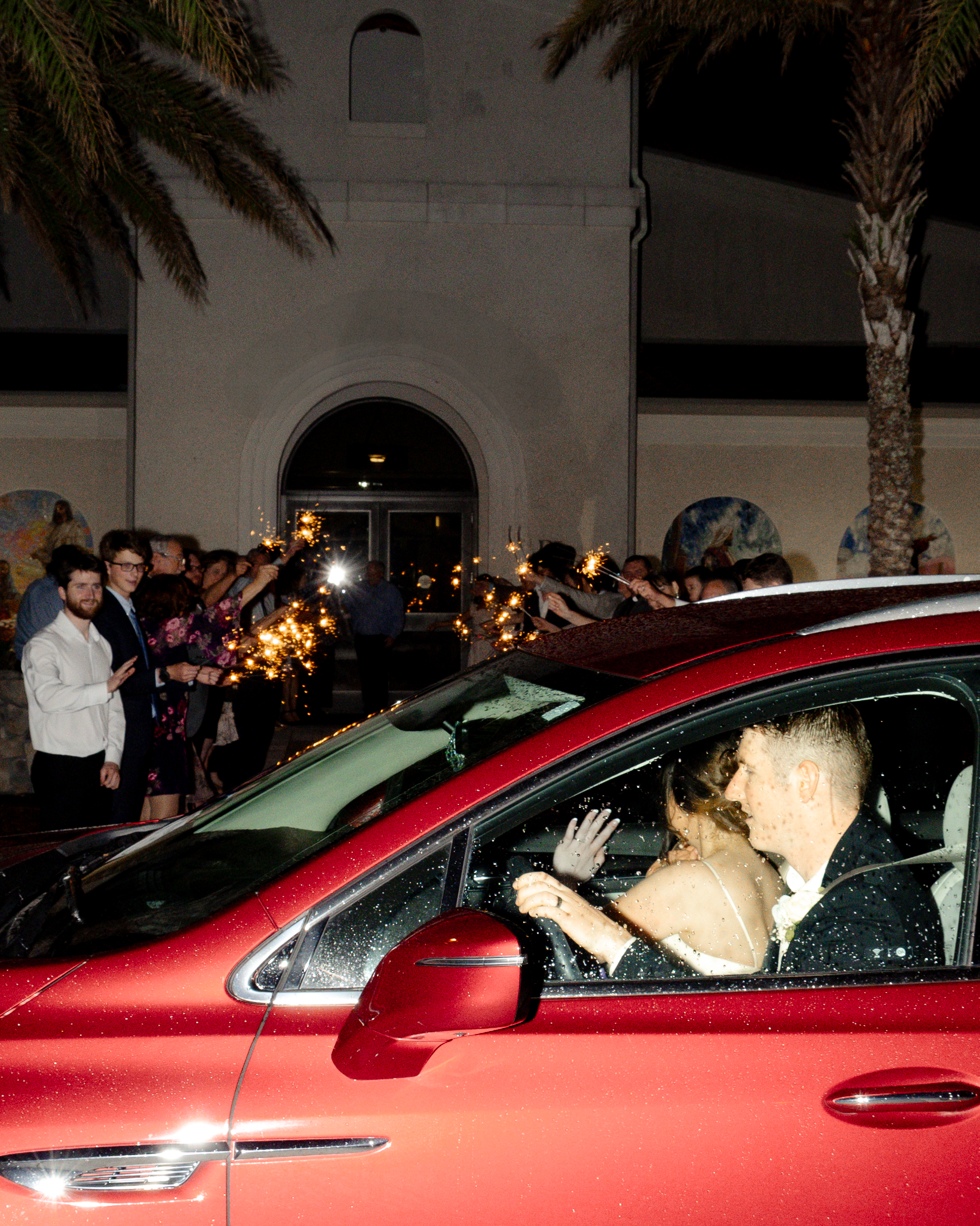 Bride and groom wave goodbye from their red getaway car as guests light sparklers outside the St. Enda of Aran Formation Center, concluding a beautiful Catholic wedding reception.
