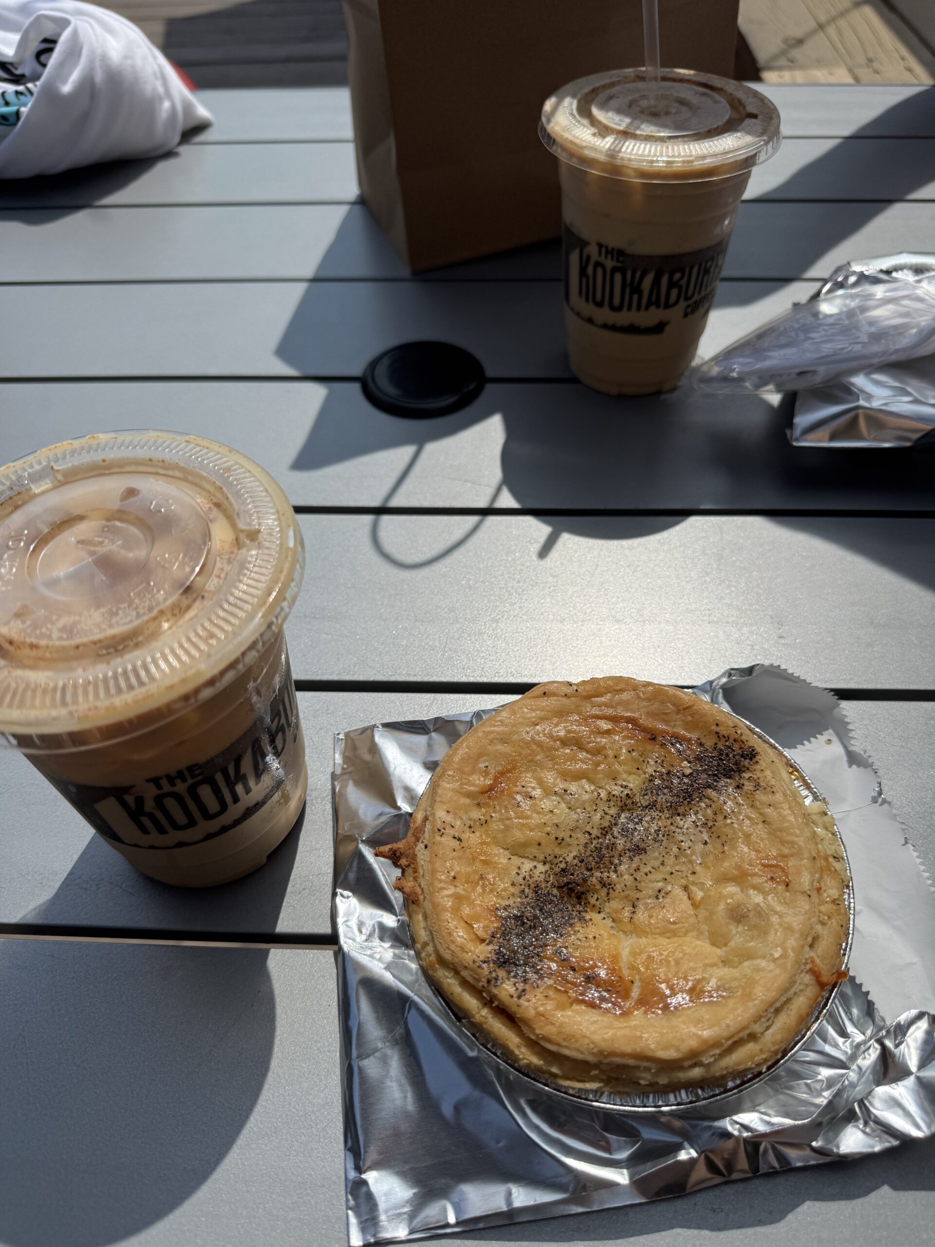 Two Catholic wedding photographers enjoying breakfast before a big wedding day at The Kookaburra café. The photo features two iced coffees and a savory meat pie on a table in the bright morning sunlight, setting the tone for a busy day ahead.