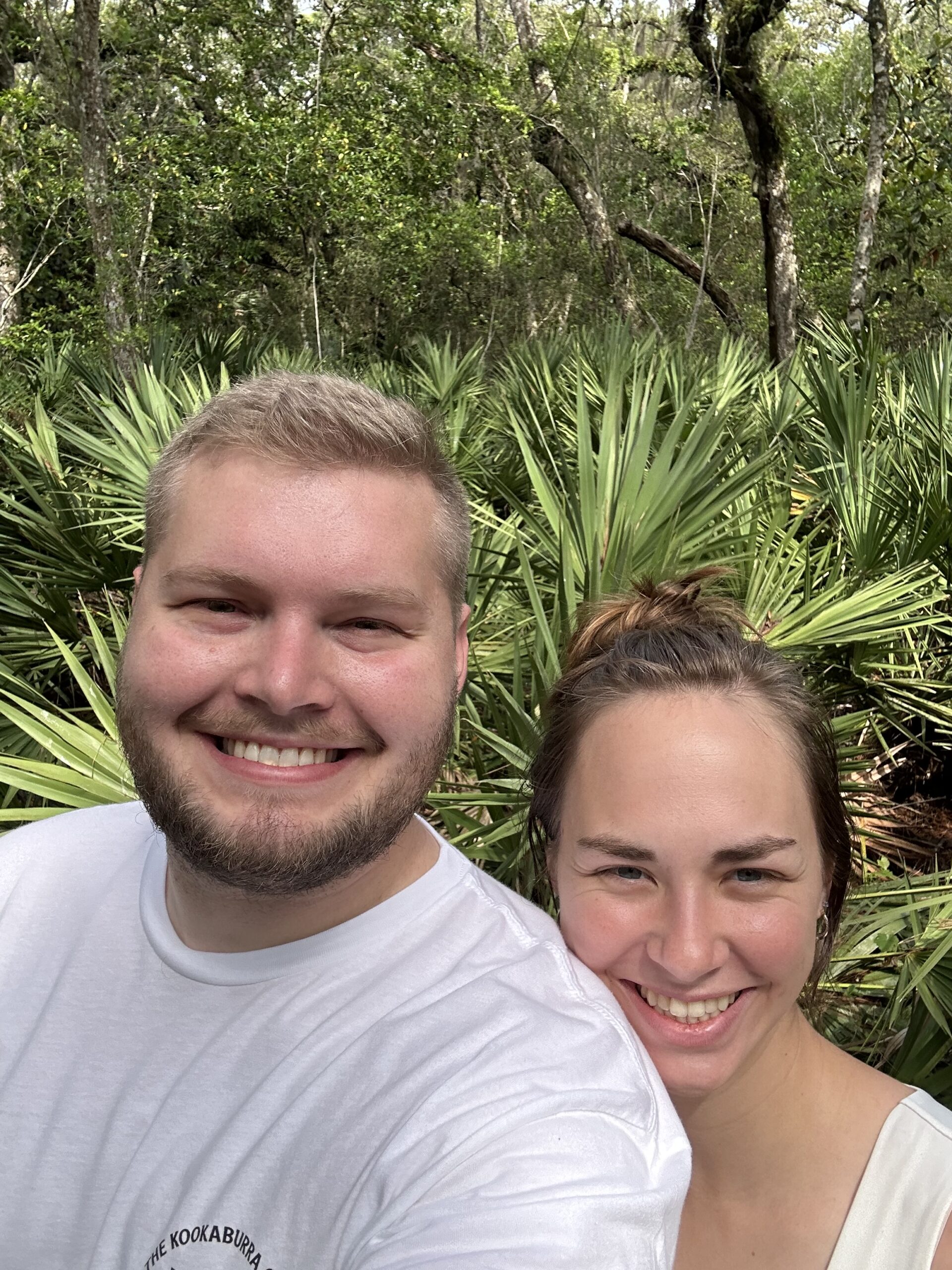 A joyful selfie of two Catholic wedding photographers, capturing a moment of relaxation before a big wedding. The couple smiles in front of lush greenery, ready for their wedding photography duties after a peaceful morning meal together.