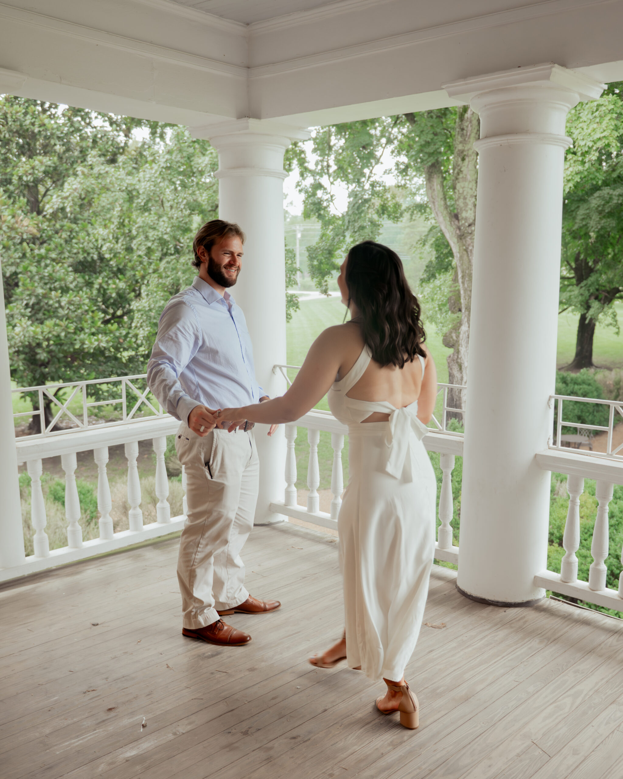 A catholic couple playfully dances with each other on the front porch of a tennessee historic home during their summer engagement session
