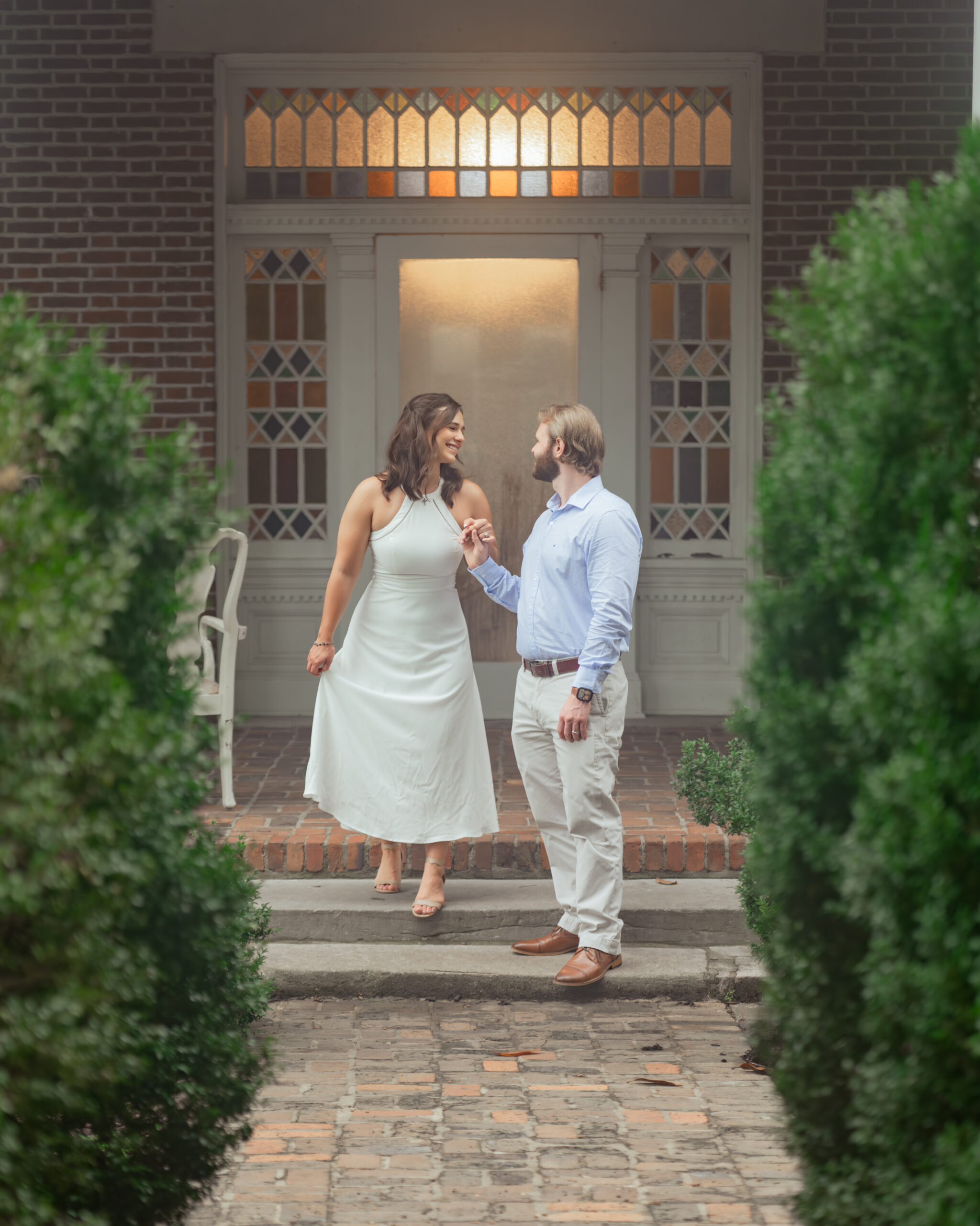A catholic groom leads his bride down the porch steps of a historic mansion in thompson's station tennessee during their hazy summer engagement session