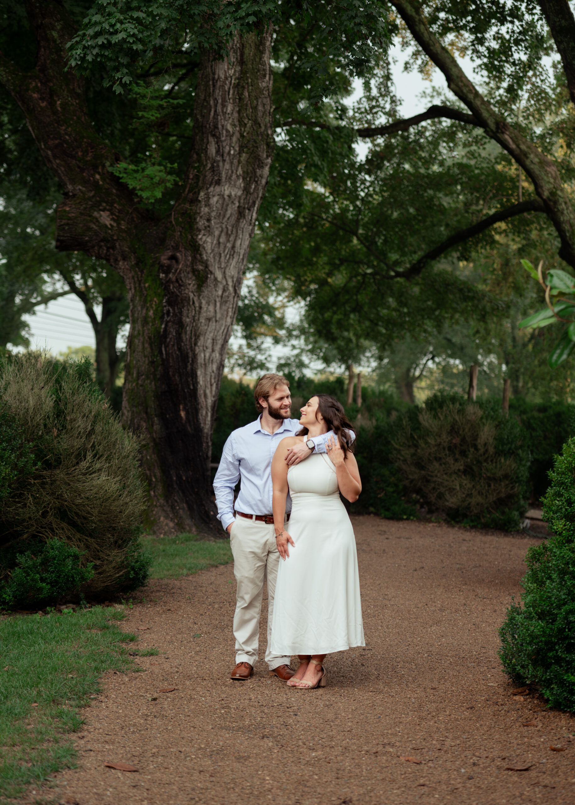 a catholic man hugs his fiancee from behind as she gazes lovingly at him in front of a large historic tree during this tennessee summer engagement session