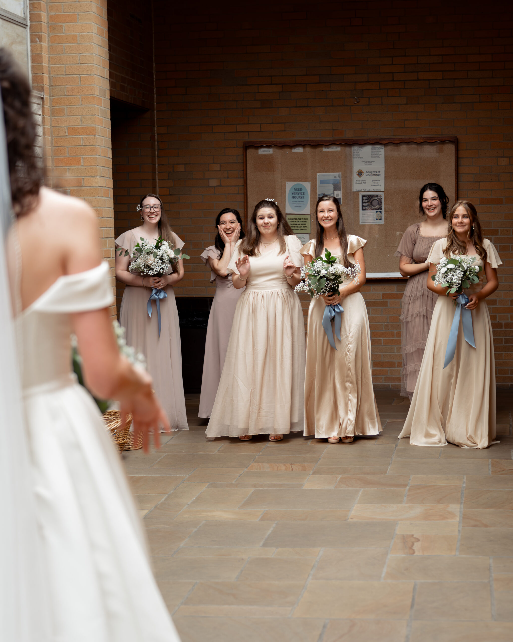 a group of bridesmaids comes around a corner of holy spirit catholic church to show their tearful and joyful surprise at seeing a catholic bride in her wedding dress