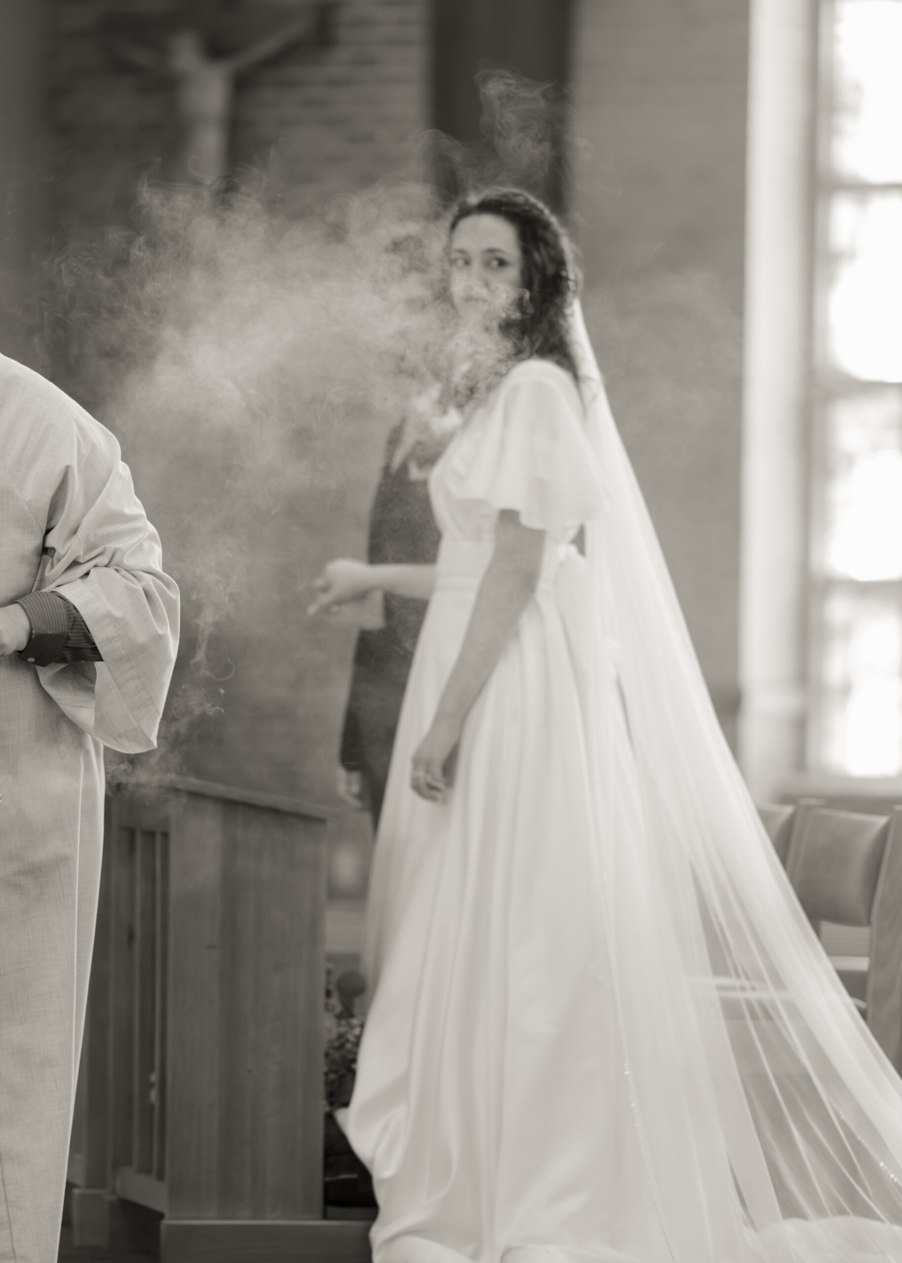 a detail photograph of incense during a catholic wedding Mass with the bride and groom standing behind it, the bride is looking back at the guests