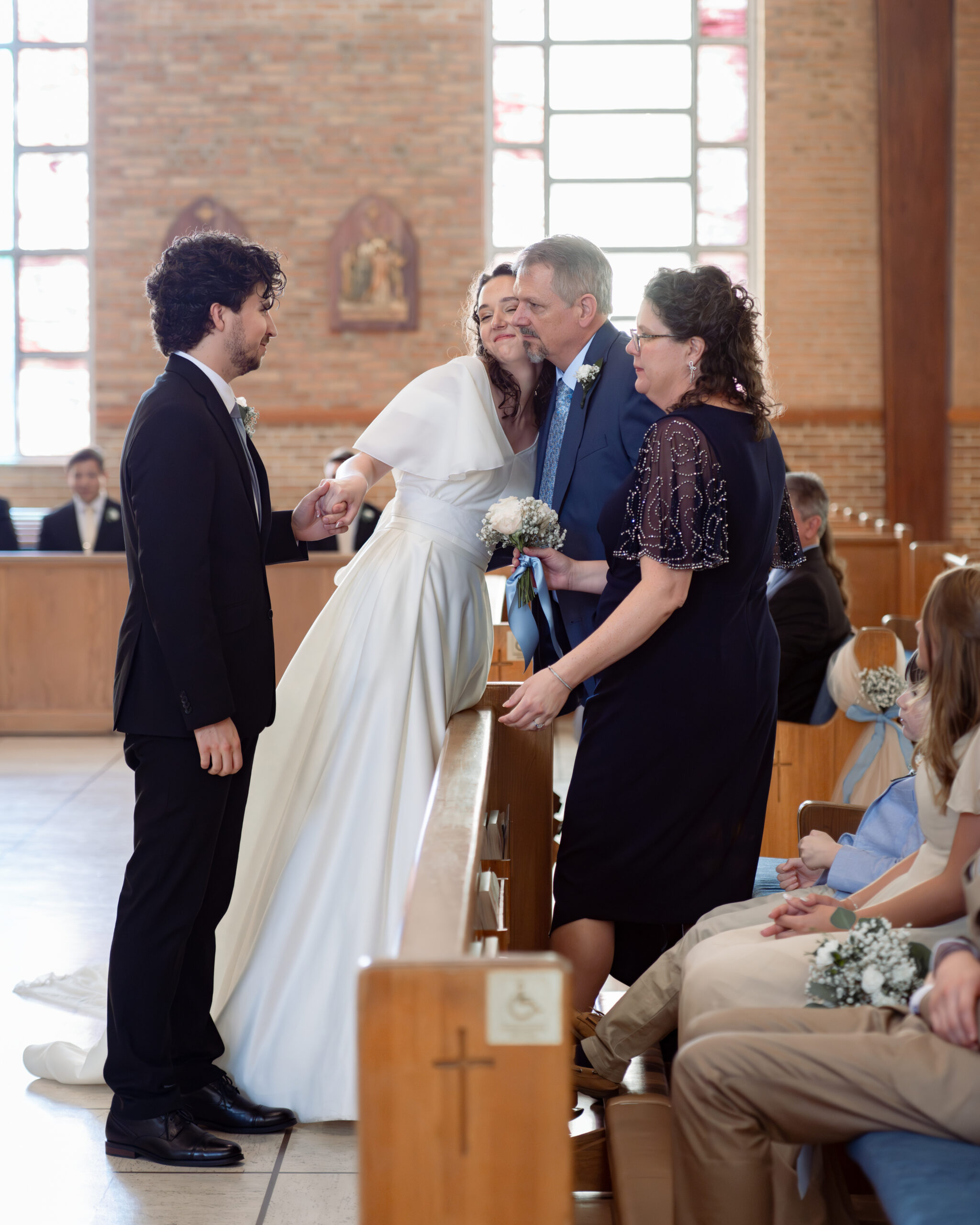 A catholic bride and groom tearfully hug the bride's parents during their catholic wedding Mass at holy spirit catholic church in huntsville, alabama