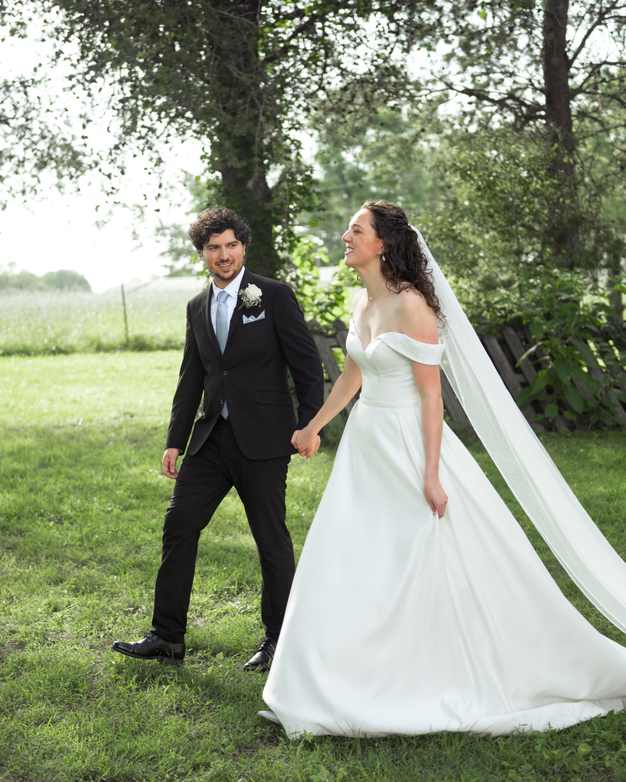 A catholic bride and groom laugh and walk surrounded by bright summer green at their reception venue in athens, alabama