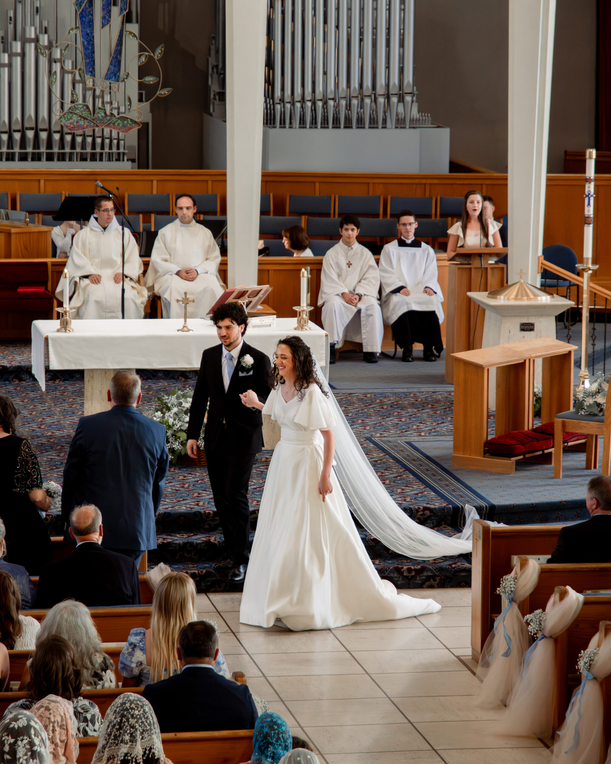 A catholic bride and groom walk over to receive a bouquet from the bride's parents during their catholic wedding Mass at holy spirit catholic church in huntsville, alabama