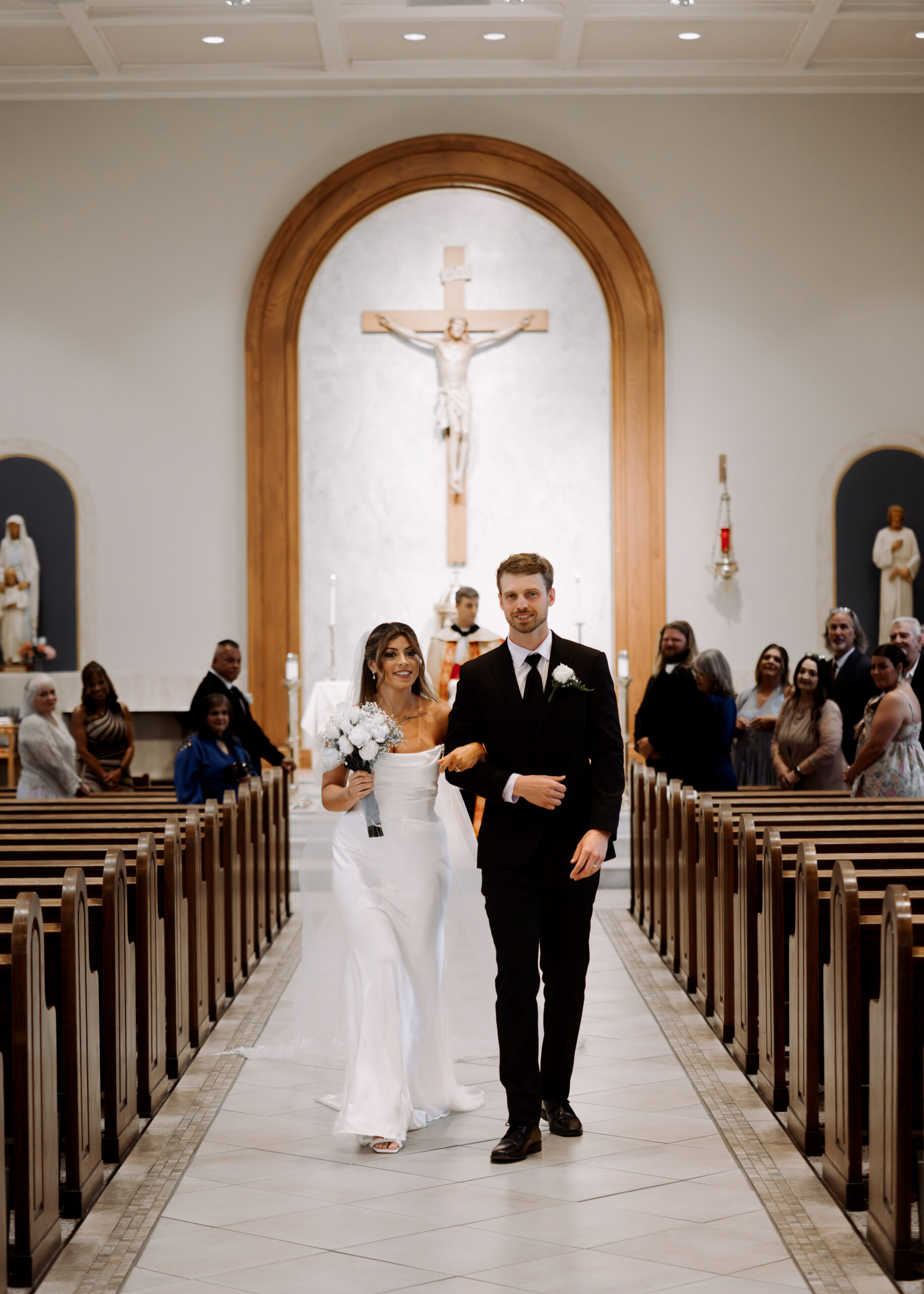 A catholic bride and groom smile as they walk up the aisle with their family looking on during a catholic wedding ceremony at St. Ann's Catholic Church in Nashville, TN