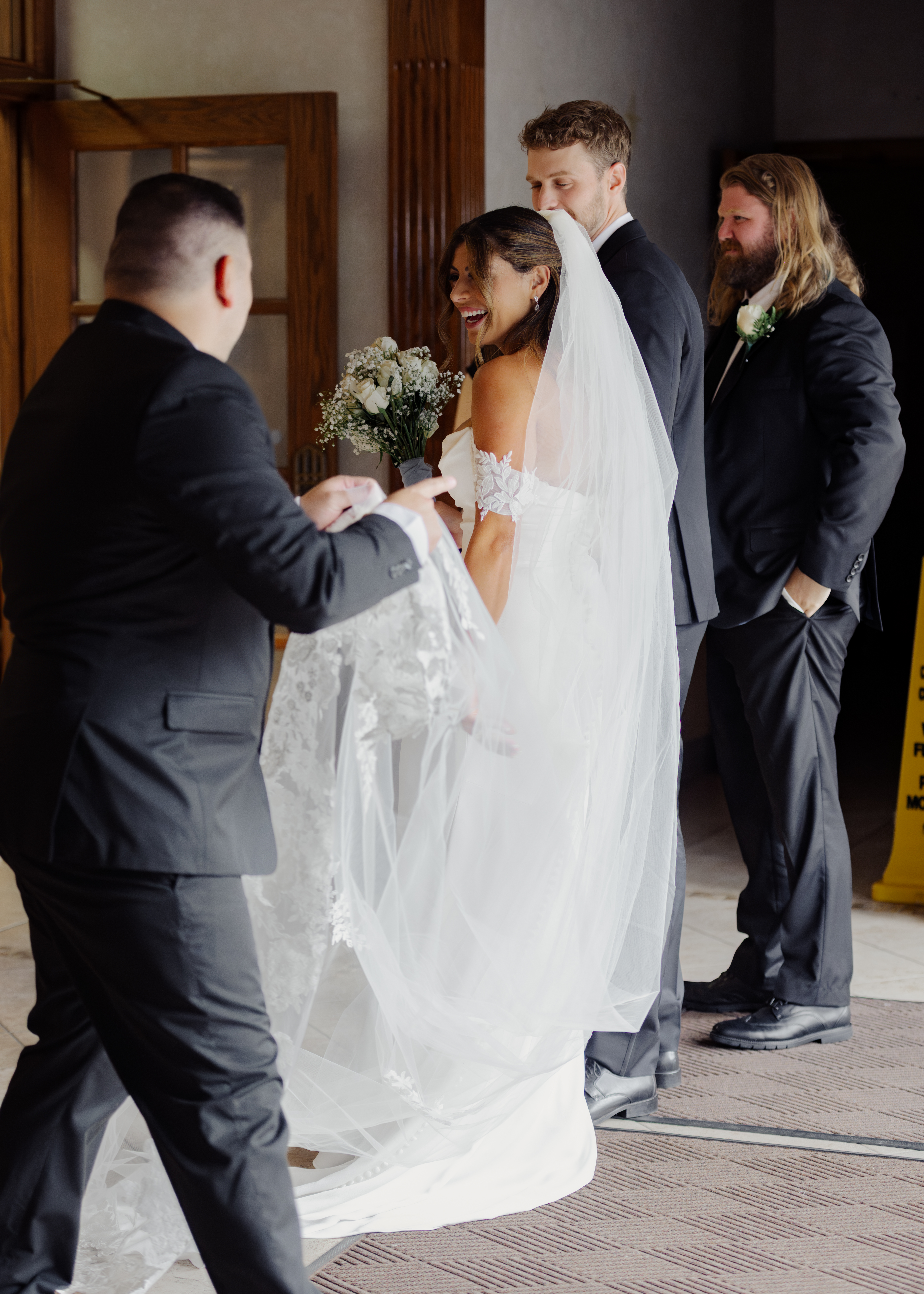 A catholic bride and groom laugh at her brother as he adjusts her train, everyone is filled with authentic joy, after a Catholic wedding ceremony at St. Ann's Catholic Church in downtown Nashville.